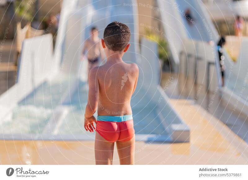 Anonymes Kind, das in der Nähe des Rutschenausgangs steht Badebekleidung ohne Hemd nackte Schultern Sommerzeit Windstille Kindheit Sliden Urlaub Junge Wasser