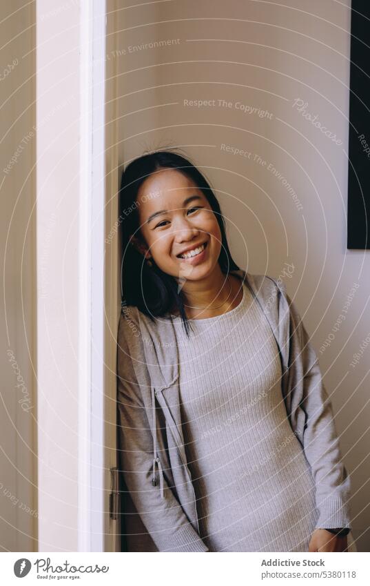 Lächelnde asiatische Frau in Freizeitkleidung Raum positiv Fenster Glück ruhen heimwärts gemütlich lässig froh ethnisch heiter jung Optimist Appartement Freude
