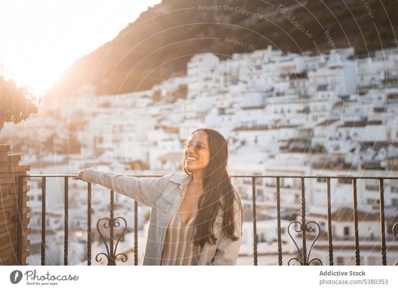 Junge Frau mit Sonnenbrille auf dem Balkon stehend Stadt Lächeln genießen Tourist Berge u. Gebirge Urlaub Zaun Gebäude jung Glück heiter Sommer Haus bewundern