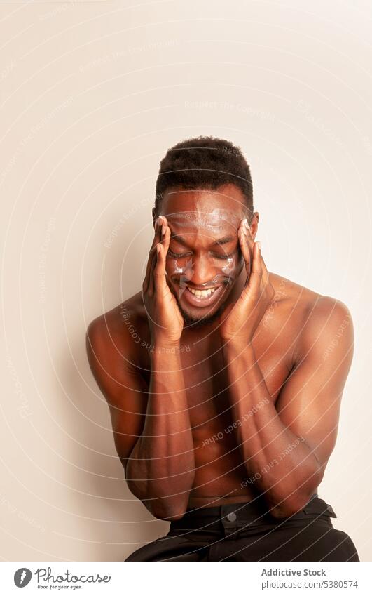 Glücklicher schwarzer Mann mit nacktem Oberkörper beim Eincremen im Studio Hautpflege Sahne bewerben Gesicht täglich Feuchtigkeit Kosmetologie Verfahren