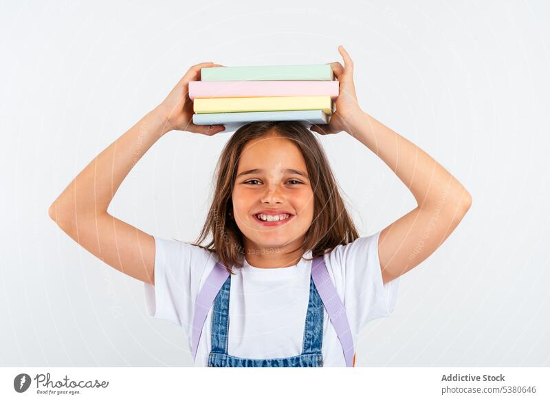 Fröhliches Schulmädchen mit Büchern auf dem Kopf Mädchen Kind Buch farbenfroh Stapel Gleichgewicht Lächeln Haufen Uniform Schule positiv Bildung Pupille Glück