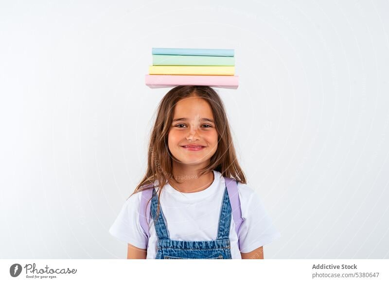 Lächelndes Schulmädchen mit Büchern auf dem Kopf Mädchen Kind Buch farbenfroh Stapel Gleichgewicht Haufen Uniform Schule positiv Bildung Pupille Glück wenig