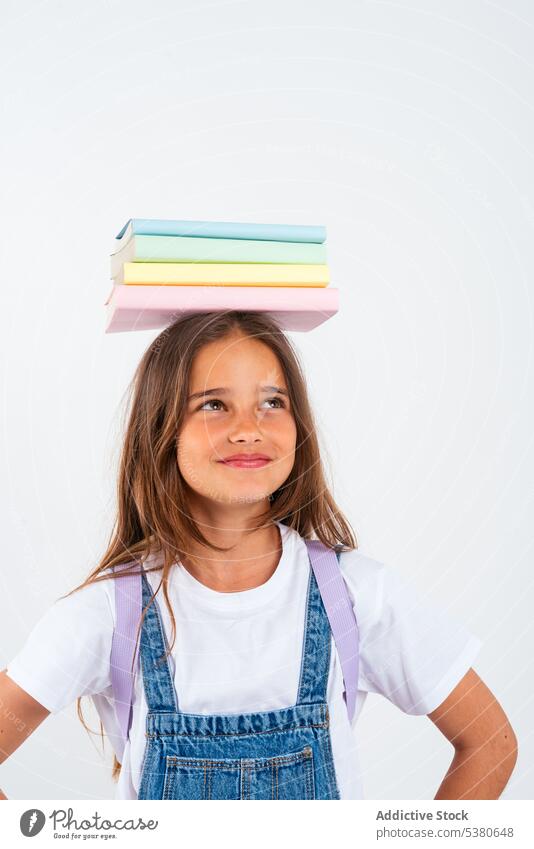 Lächelndes Schulmädchen mit Büchern auf dem Kopf Mädchen Kind Buch farbenfroh Stapel Gleichgewicht Haufen Uniform Schule positiv Bildung Pupille Glück wenig