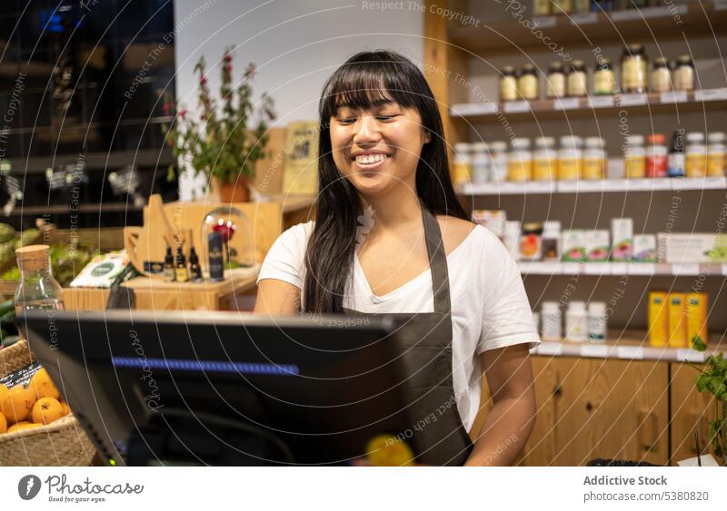 Fröhliche asiatische Frau am Schalter mit Computer Verkäufer Werkstatt Abfertigungsschalter Supermarkt Arbeit Kassierer benutzend positiv Lebensmittel Schürze