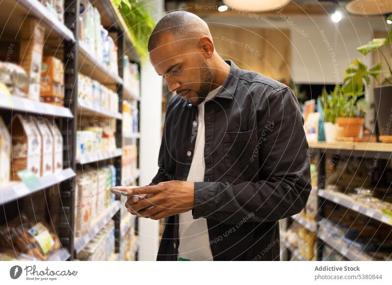 Konzentrierter schwarzer Mann benutzt Smartphone im Lebensmittelgeschäft Werkstatt Kunde Supermarkt Kauf wählen Nachricht kaufen männlich Laden Afroamerikaner