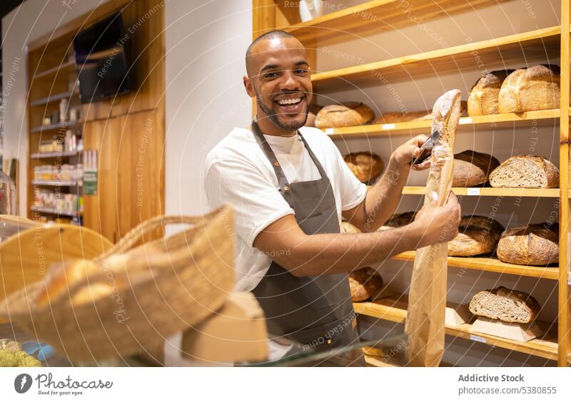 Fröhlicher schwarzer Mann packt Brot in Papiertüte Bäckerei Verkäufer Tüte Lebensmittelgeschäft Werkstatt heiter Kleinunternehmen Arbeit gebacken Besitzer