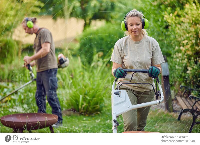 Junge Gärtnerin, die einen Rasenmäher benutzt, während ihr Kollege oder Chef im Hintergrund den Garten trimmt Kunstgewerbler Bauherr Kettensäge Konzept