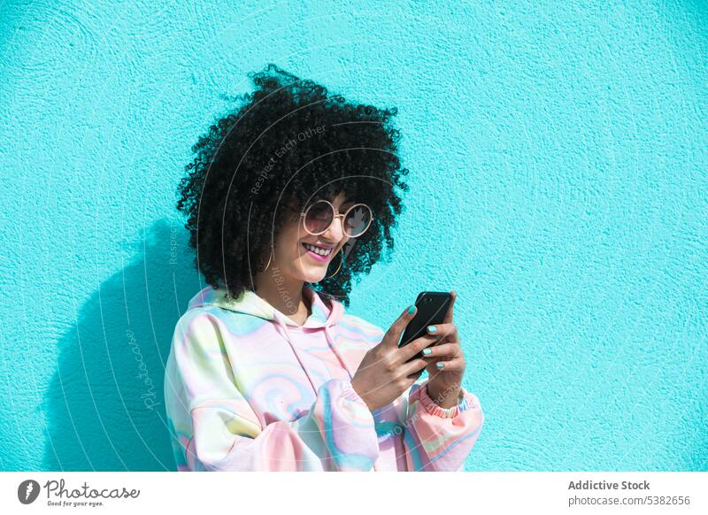 Lächelnde ethnische Frau mit Sonnenbrille schreibt Texte auf Smartphone benutzend Glück Nachricht Textnachricht Internet Sommer jung Funktelefon Straße Mobile