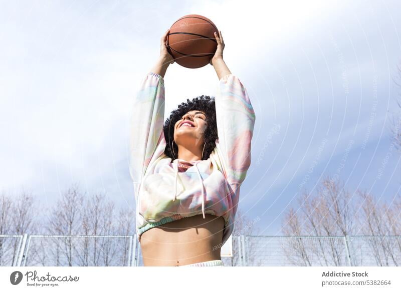Fröhliche ethnische Frau hält Basketball in ausgestreckten Händen Augen geschlossen Blauer Himmel Spieler Lächeln Arm angehoben Gericht positiv Sommer jung