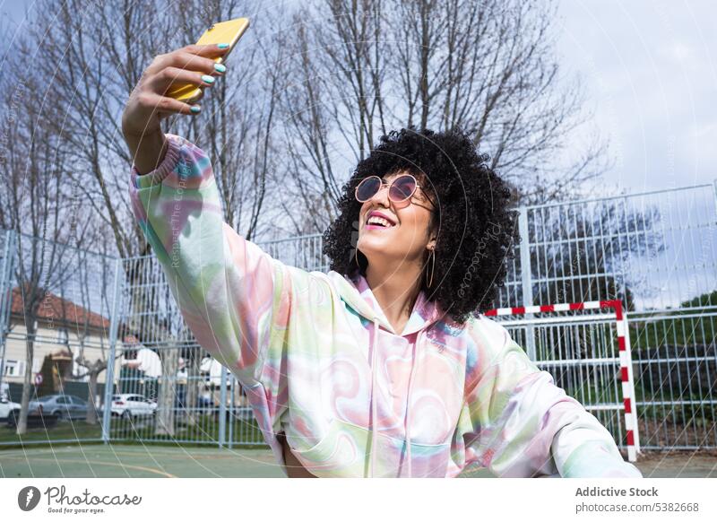 Fröhliche ethnische Frau macht Selfie mit Smartphone Glück heiter Sportpark Zaun Selbstportrait Moment Sommerzeit jung hispanisch positiv Sonnenlicht Mobile