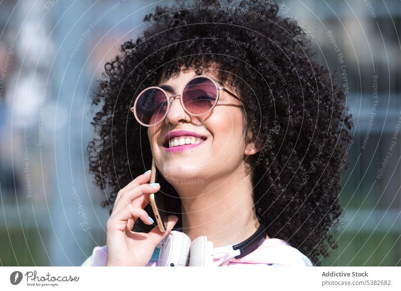 Fröhliche ethnische Frau steht und spricht mit einem Smartphone Kopfhörer reden sprechen heiter Telefonanruf Lächeln positiv Gespräch jung hispanisch Straße