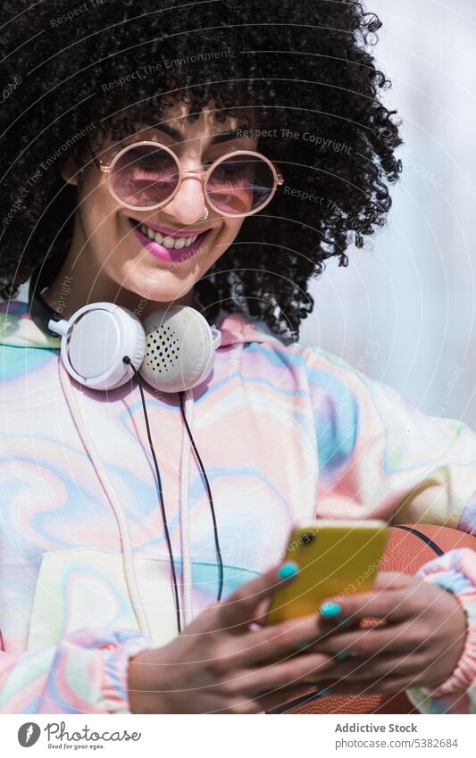 Glückliche ethnische Frau mit Sonnenbrille beim Surfen auf dem Smartphone benutzend Textnachricht Lächeln positiv Kopfhörer lesen Internet online jung Browsen