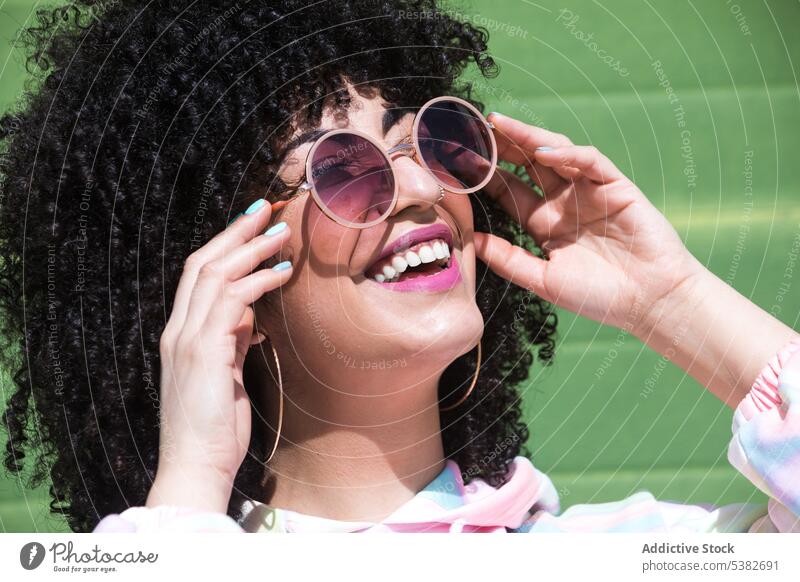 Selbstbewusste ethnische Frau in trendigem Outfit steht in der Nähe der grünen Wand Sportpark Sonnenbrille Frisur Vorschein Mode selbstbewusst Sonnenlicht
