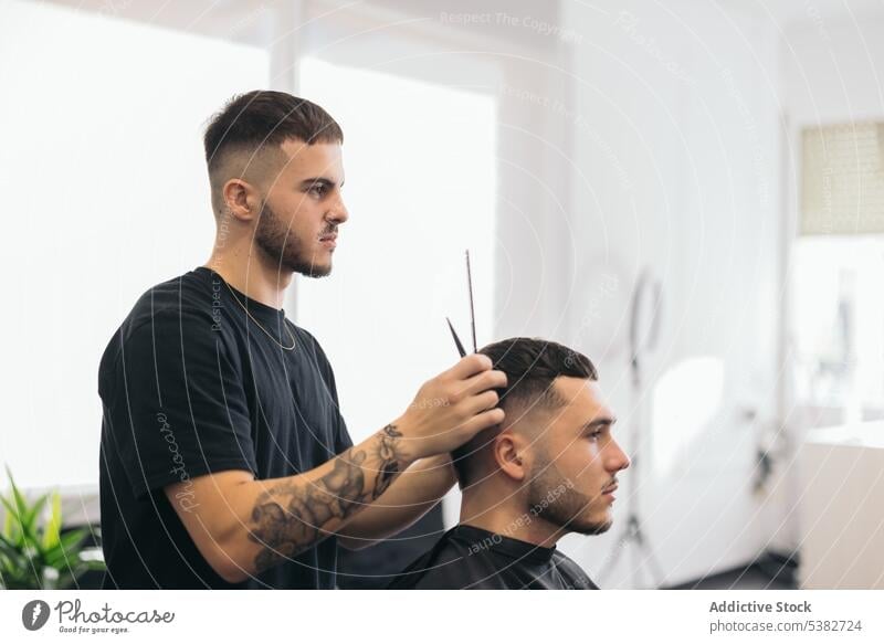 Junger Friseur schneidet Haare mit einer Schere im Friseursalon Barbershop Vollbart bärtig Bartiger Mann Schönheit Klinge Pflege Klient Ausschnitt Frisur Kamm