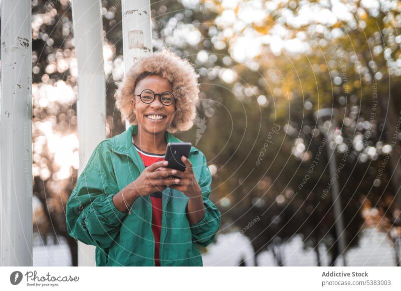 Fröhliche junge Frau benutzt ihr Smartphone im Freien heiter benutzend Mobile Telefon positiv Browsen Lächeln Park Brille schwarz Glück Nachricht Afroamerikaner