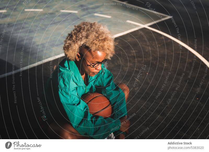 Schwarze Frau mit lockigem Haar sitzt mit Basketball auf dem Boden Sportlerin Spieler Sportpark Ball Abend Sportbekleidung sitzen krause Haare ruhen Gericht