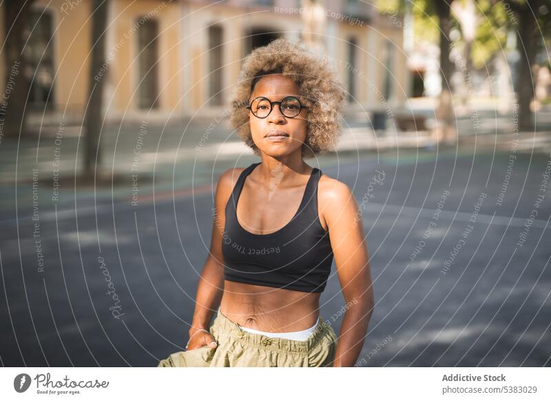 Seriöse schwarze Frau in Sport-BH und Brille auf der Straße stehend heiter krause Haare sportlich urban Großstadt Afroamerikaner jung ethnisch tagsüber