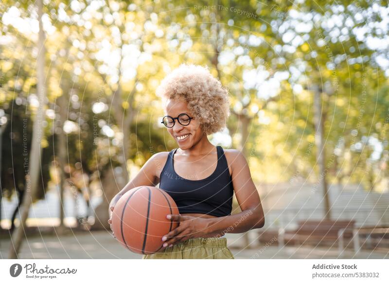 Fröhliche junge Frau hält lächelnd einen Basketball im Park Spieler Sport Ball Sportpark Athlet Sportlerin Afroamerikaner schwarz spielen Gericht Inhalt