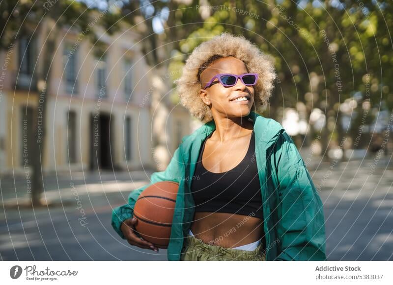 Lächelnde Frau mit modischer Sonnenbrille und Basketball auf der Straße Porträt Stil trendy Glück positiv selbstbewusst Großstadt heiter froh Sommer modern jung