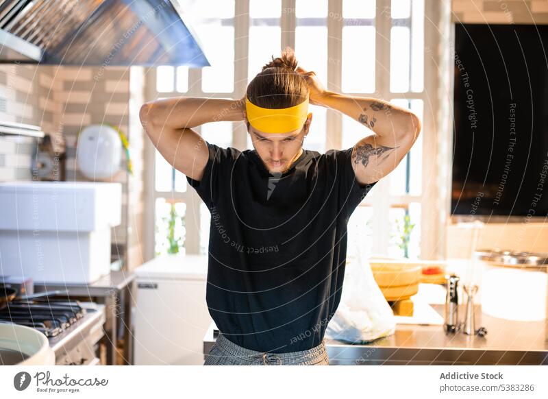 Konzentrierter junger Mann beim Binden des Stirnbands in der Küche vor dem Kochen Fokus Küchenchef Krawatte ernst Konzentration Utensil Küchengeräte männlich