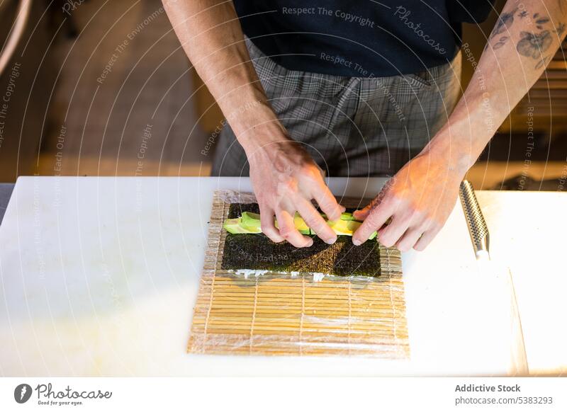 Unbekannter Mann arrangiert Avocadoscheiben auf Seetangwickel rollen anonym Tisch Küche vorbereiten Bestandteil Lebensmittel Bambus umhüllen Seegras männlich