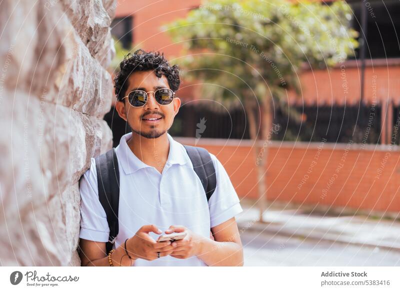 Positiv eingestellter Mann auf der Straße Steinwand Sommer Stil zufrieden urban Lächeln Sonnenlicht Urlaub männlich Großstadt hispanisch Typ Sonnenbrille Wand