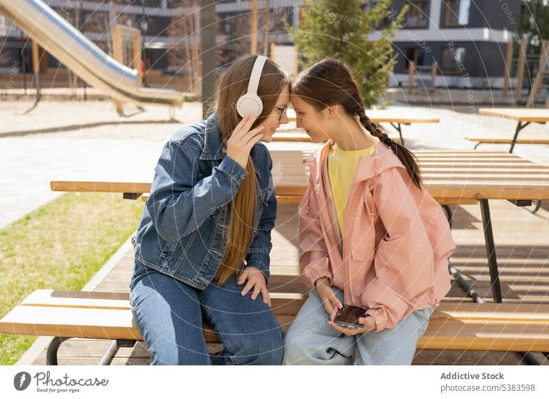 Inhalt: Teenager-Freunde verbringen die Sommerzeit im Park Mädchen Zusammensein Freizeit Stirn berühren Bonden benutzend Smartphone Kopfhörer Bank jung positiv