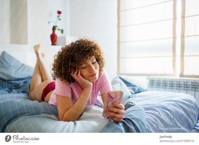 Junge verliebte Frau mit lockigem Haar, die sich etwas Schönes auf ihrem Smartphone ansieht benutzend soziale Netzwerke Lächeln Nachricht ruhen Komfort Bett