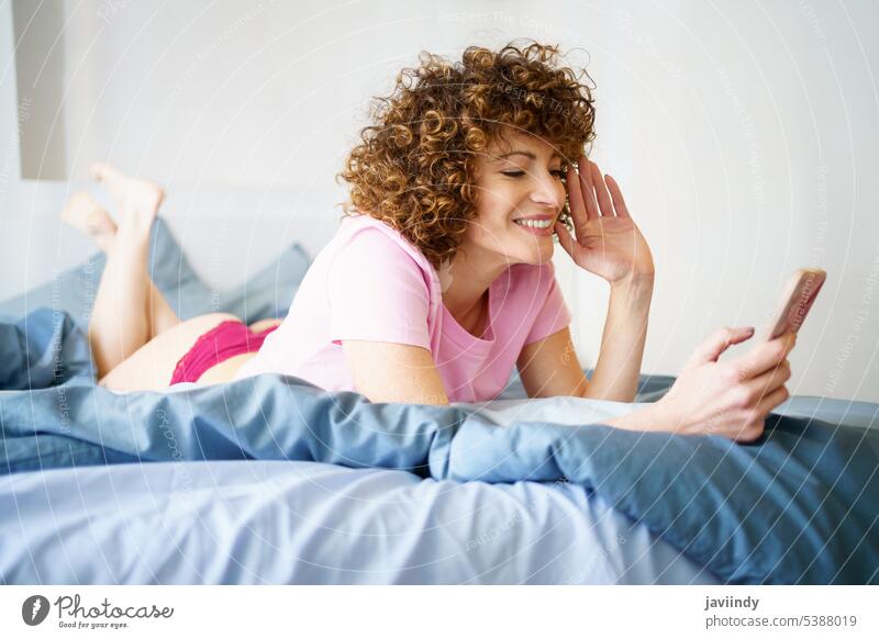 Lächelnde Frau, die sich im Bett per Videochat über ihr Smartphone unterhält Wellenhand Videoanruf benutzend Gruß Anschluss soziale Netzwerke heimwärts Glück