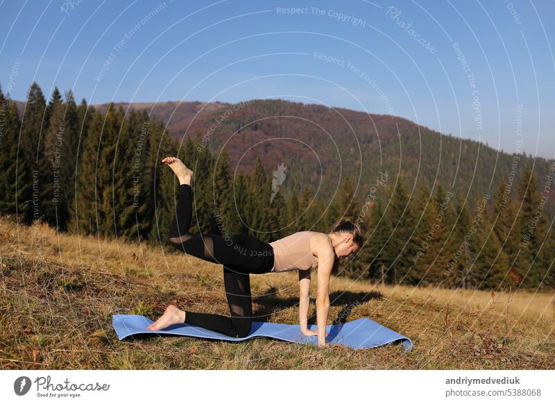 Junge fitte Frau macht Übungen auf der Yogamatte auf dem Hintergrund der sonnigen Berge Hügel. Outdoor-Workout, gesunder Lebensstil. Frau im Sportanzug macht Asanas in der Natur gegen den Wald am Morgen