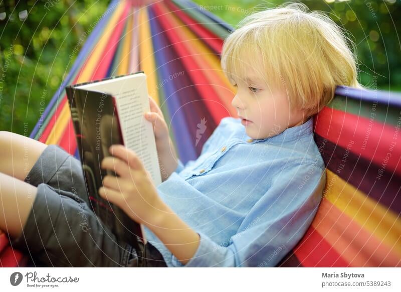 Netter kleiner blonder kaukasischer Junge, der ein Buch liest und Spaß mit einer bunten Hängematte im Hinterhof oder auf einem Spielplatz hat. Sommerfreizeit für Kinder. Kind schwingt und entspannt in der Hängematte.