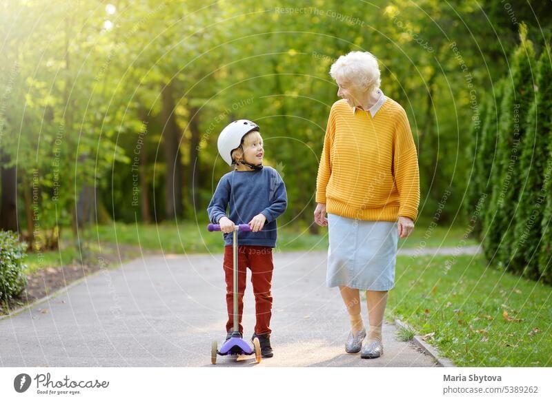 Schöne Oma und ihr kleines Enkelkind gehen zusammen im Herbst Park. Junge fährt mit dem Motorroller. Kind Spaziergang Holunderbusch Großmutter Familie