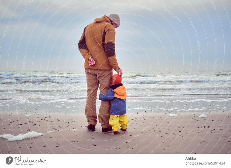 Ein Kleinkind hält sich an einem Mann fest während er es beruhigend am Kopf krault, Strand, im Hintergrund Wellen Vater mit Kind Vertrauen Zuneigung