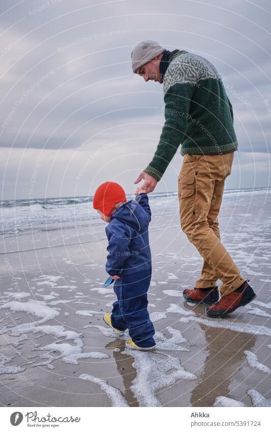 Ein Mann im Strickpullover und heller Hose hilft einem kleinen Kind mit roter Mütze bei der Erkundung am Strand erste Schritte Kleinkind Vater Vertrauen
