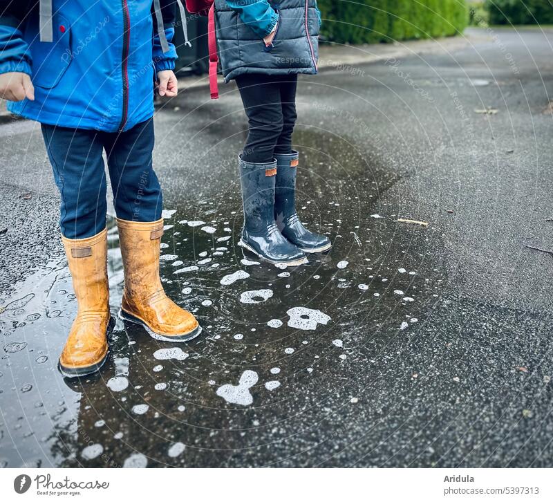 Zwei Kinder stehen mit Gummistiefeln in einer Pfütze nass Asphalt Außenaufnahme Regen Spielen Kindheit Wetter schlechtes Wetter Wasser Freude Herbst