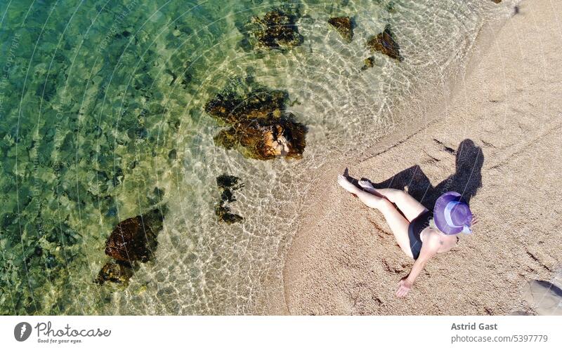 Eine Frau mit Badeanzug und Strohhut sitzt im Sand am Meer luftaufnahme drohnenfoto frau seniorin urlaub meer italien strand sandstrand wasser steine felsen