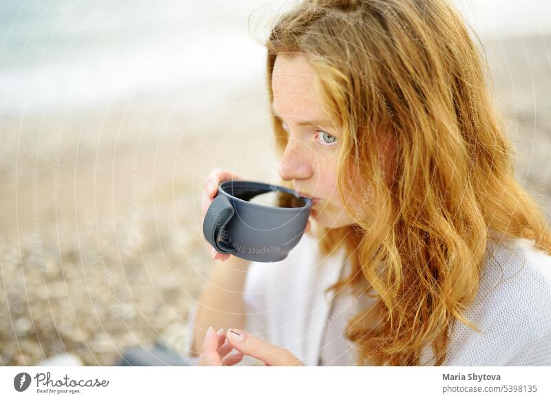 Eine junge rothaarige Frau macht ein Picknick am Meeresufer. heiß trinken Thermoskanne Tee Getränk MEER Erwachsener Strand Familie Person herumhängen Ufer