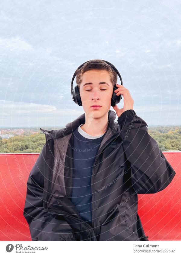 Teenager im Riesenrad mit Kopfhörern Musik Musik hören Freude Technik & Technologie Jugend Lifestyle Junge lächeln brünett Bäume am Horizont rot Regenjacke