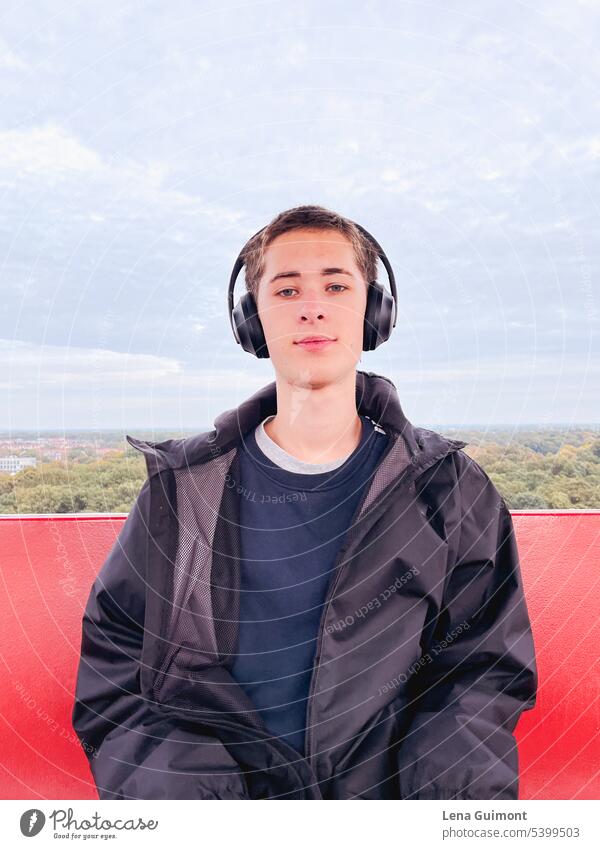 Teenager im Riesenrad mit Kopfhörern Musik Musik hören Freude Technik & Technologie Jugend Lifestyle Junge lächeln brünett Bäume am Horizont rot Regenjacke