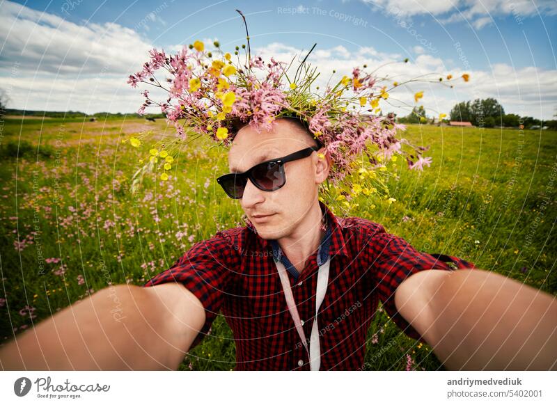Junger lächelnder Mann mit Wildblumenkranz und Sonnenbrille macht ein Selfie mit einer Weitwinkelkamera und hat Spaß auf dem Feld an einem Sommertag. Positive Kerl hat keine Allergie. Lifestyle Sommer Konzept.