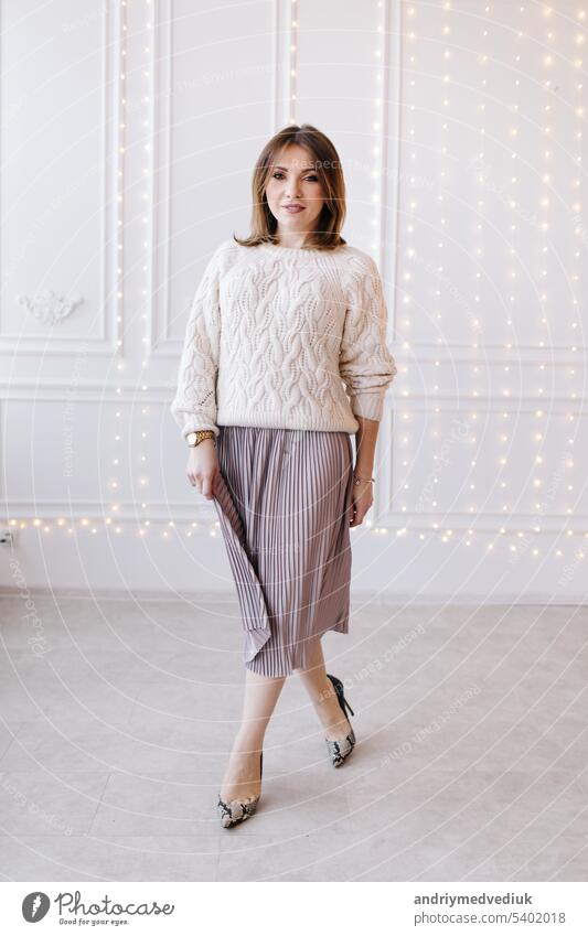 schöne junge Frau in einem weißen Pullover, Rock und High Heels auf einem Hintergrund aus weißer Wand und Weihnachtsgirlanden Mode posierend Atelier modern