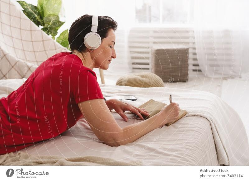 Frau hört Musik und schreibt Notizen auf dem Bett Fokus Konzentration schreiben Notebook zur Kenntnis nehmen zuhören Kopfhörer Schlafzimmer Lügen Laptop Gesang