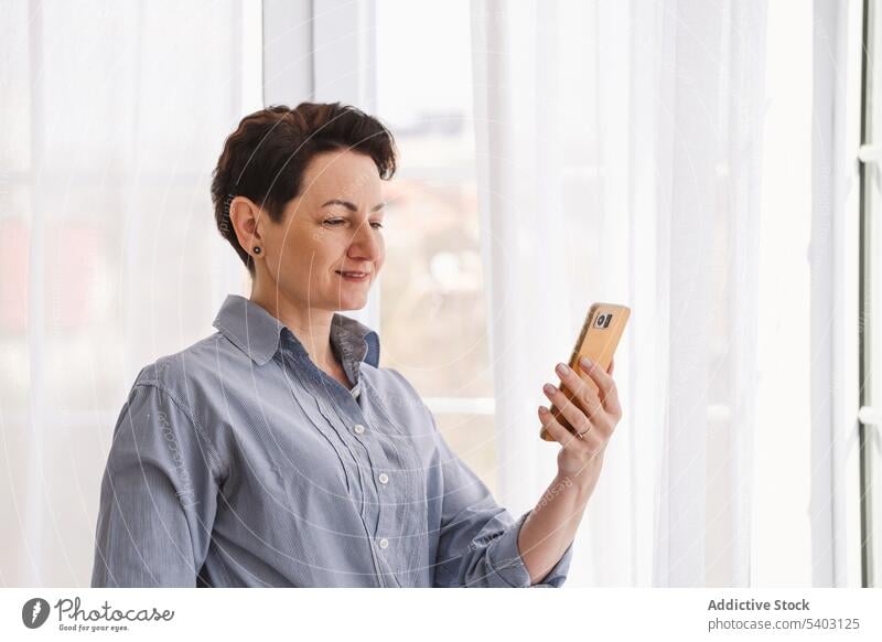 Fröhliche reife Frau beim Fotografieren mit dem Smartphone Selfie fotografieren Selbst Apparatur Gerät zu Hause Wochenende benutzend Fenster heimwärts Atelier