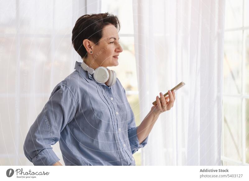 Reife Frau hält Smartphone und schaut weg Kopfhörer Apparatur Gerät zu Hause Wochenende benutzend Fenster heimwärts Atelier Stil Kurze Haare Lebensmitte Mobile