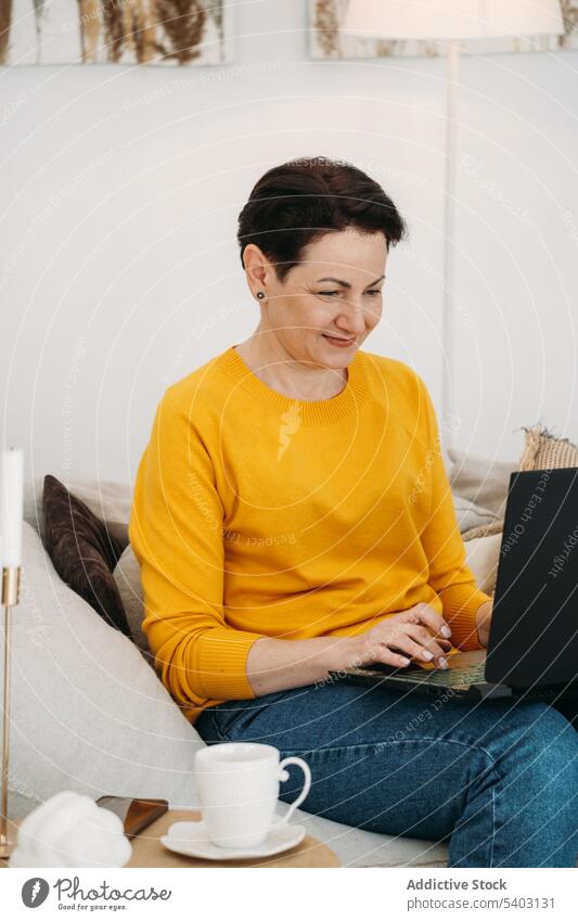 Lächelnde reife Frau arbeitet von zu Hause aus am Laptop positiv Arbeit freiberuflich Sofa Apparatur online Kaffee benutzend abgelegen heiter Gerät trinken