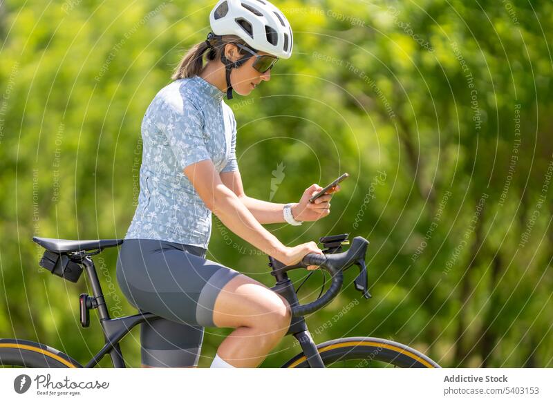 Frau auf Fahrrad mit Smartphone gegen verschwommenen Park Radfahrer Schutzhelm Sonnenbrille Sportbekleidung Mobile Browsen Athlet Rennfahrer Sicherheit