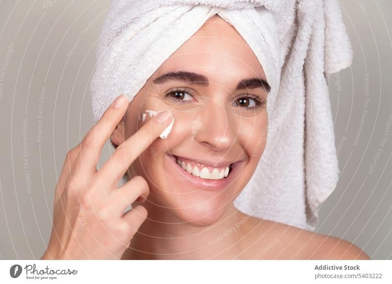 Lächelnde Frau beim Auftragen von Kosmetikcreme auf das Gesicht Hautpflege Sahne bewerben Feuchtigkeit Handtuch Leckerbissen Finger nackte Schultern Hydrat jung