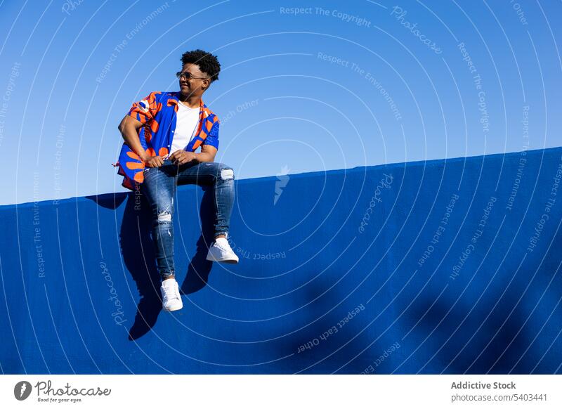 Stilvoller schwarzer Mann sitzt auf einem Zaun trendy selbstbewusst Beton Wand cool Outfit sich[Akk] entspannen Mode Turnschuh jung männlich Afroamerikaner
