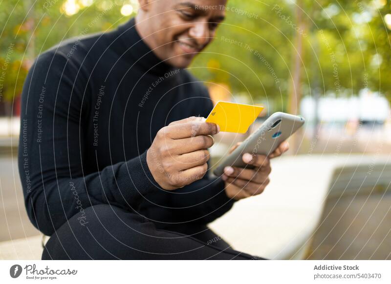Fröhlicher ethnischer Mann mit Kreditkarte und Smartphone Zahlung bezahlen positiv Kauf Glück krause Haare Park Lächeln benutzend online Apparatur Bank männlich