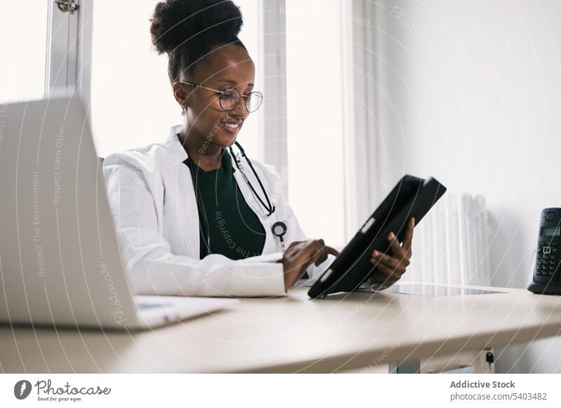 Lächelnde schwarze Frau, die am Tisch sitzt und auf einem Tablet surft Arzt Tablette benutzend Uniform Stethoskop Spezialist professionell Raum Afroamerikaner